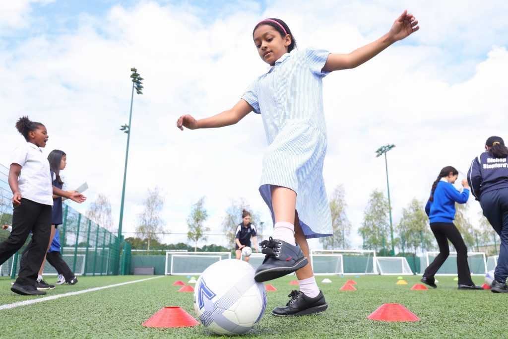 Photo of young girl kicking a football on a football pitch while wearing school uniform. There are other school children behind her.