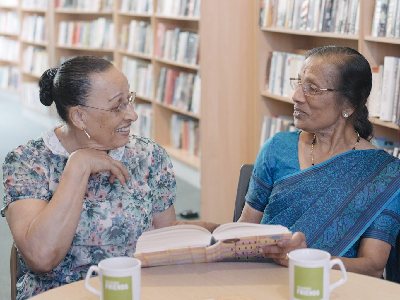 Two older woman smile as they talk and look at a book together.