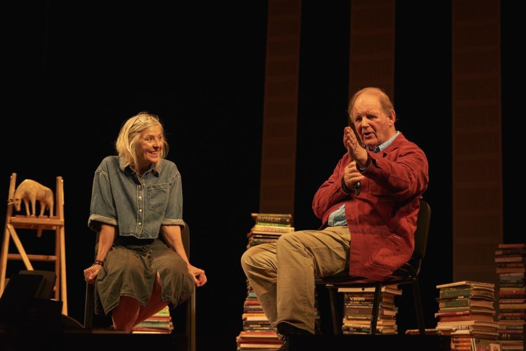 Photo of Danyah Miller and Michael Morpurgo sat on a stage with stacks of books behind them. 