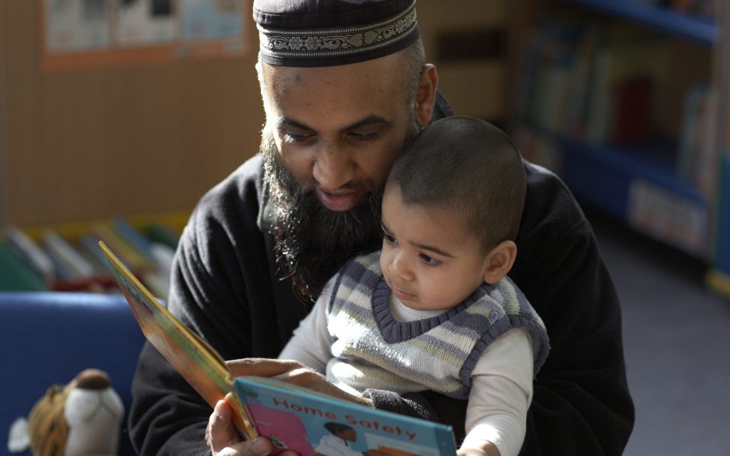 A photo of an Asian man reading a small child a book. There are bookshelves behind them.