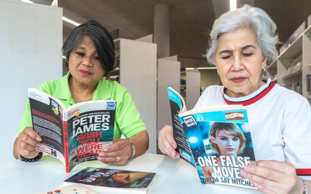 Photograph of two older Asian women sat at a table reading Quick Reads books. 