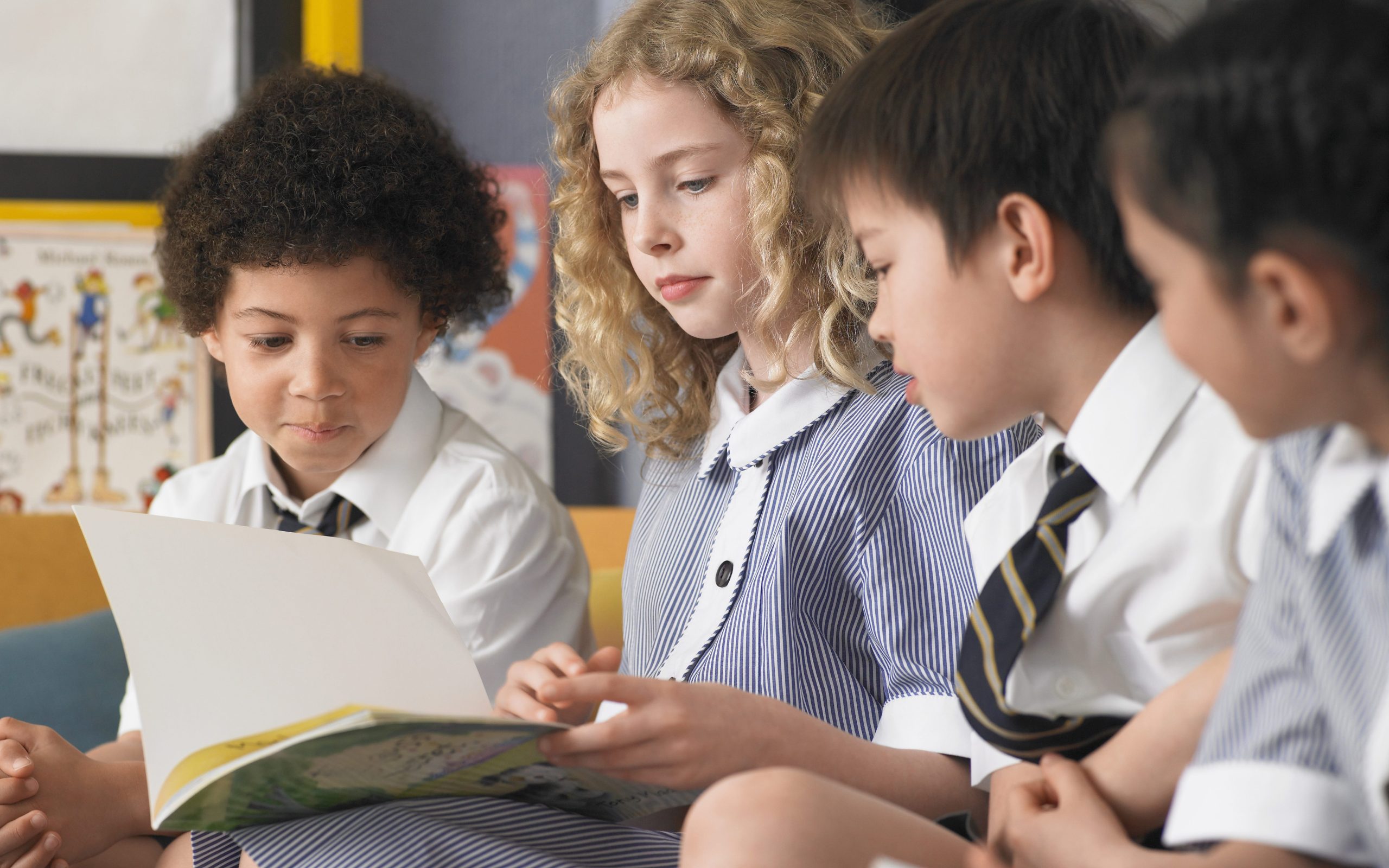 Four children sat looking at a book and wearing school uniform.