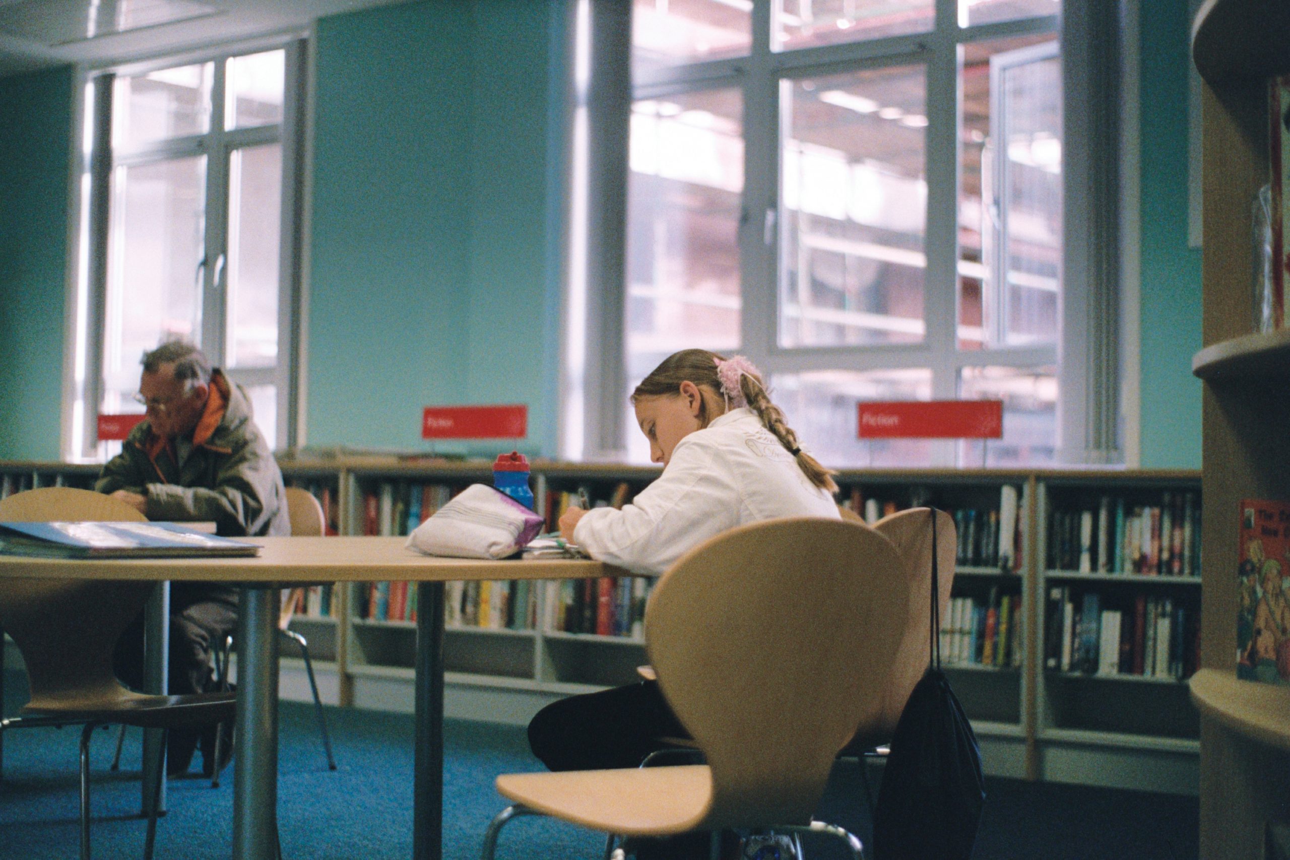 A young girl sits at a table in a library while writing.