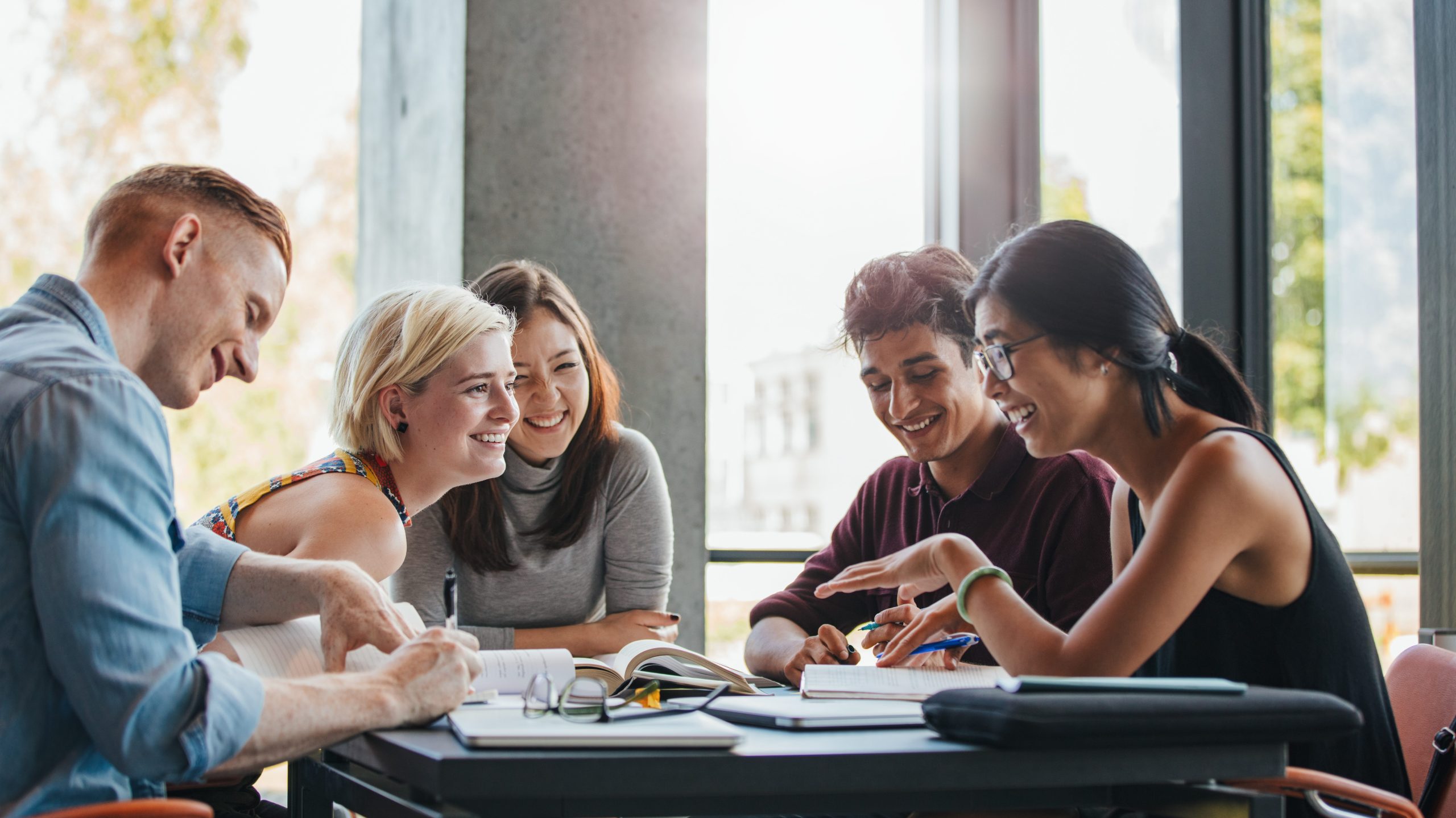 Photo of five young people sat at a table smiling and laughing. There are books on the table.