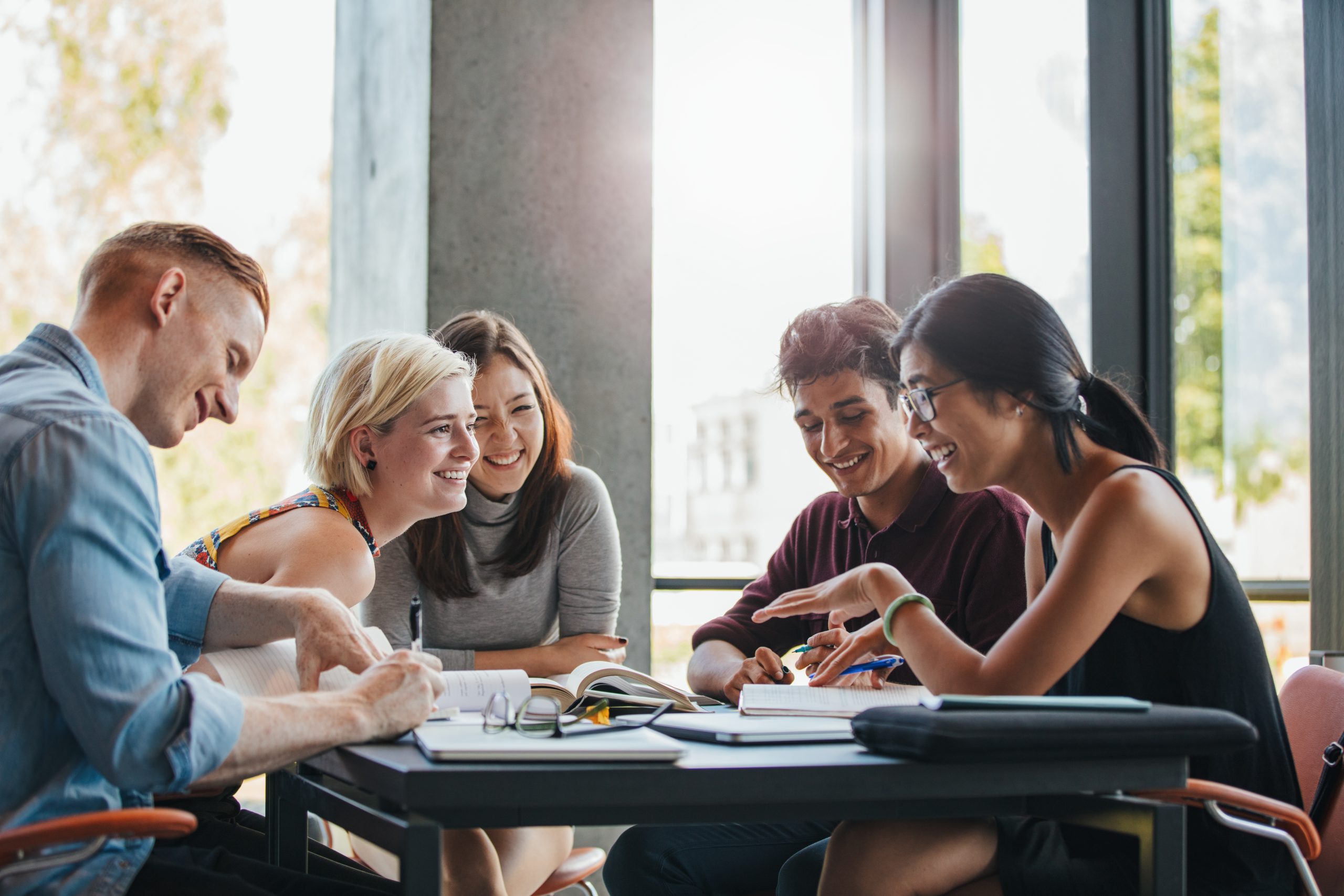 Photo of five young people sat at a table smiling and laughing. There are books on the table.