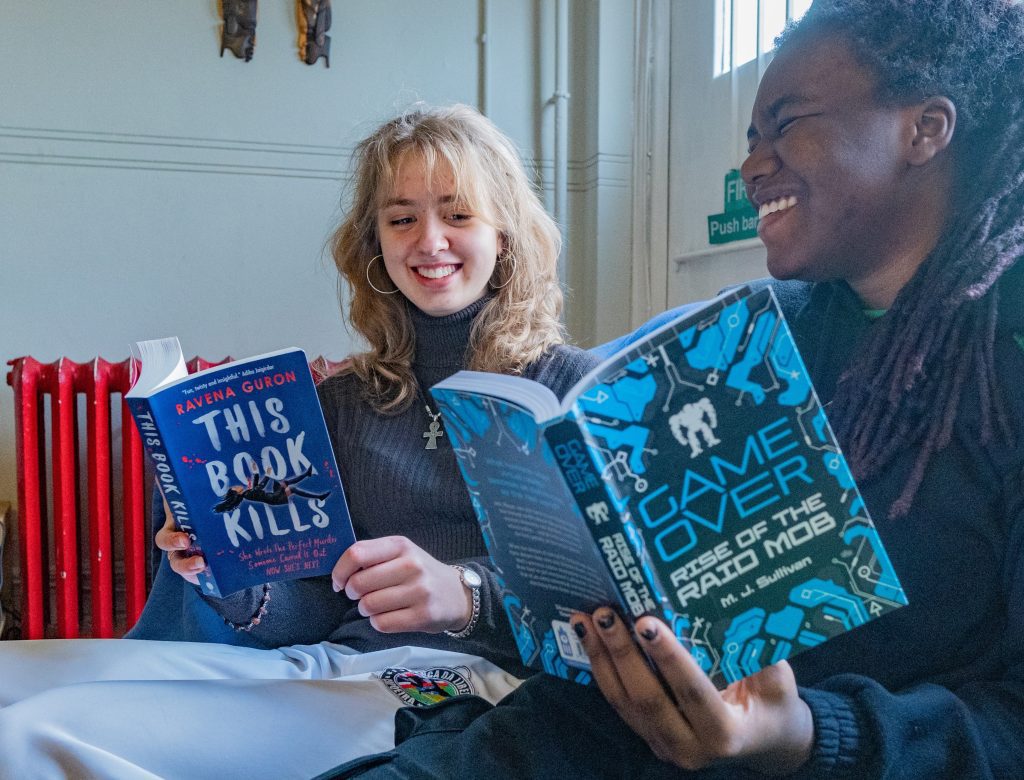 Two young women sit in a library laughing together holding copies of 'This Book Kills' and 'Game Over'