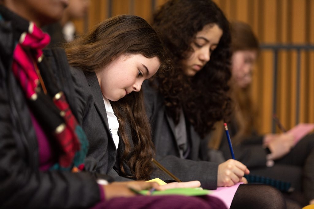 A young girl writes on a piece of paper. There are other girls around her doing the same.