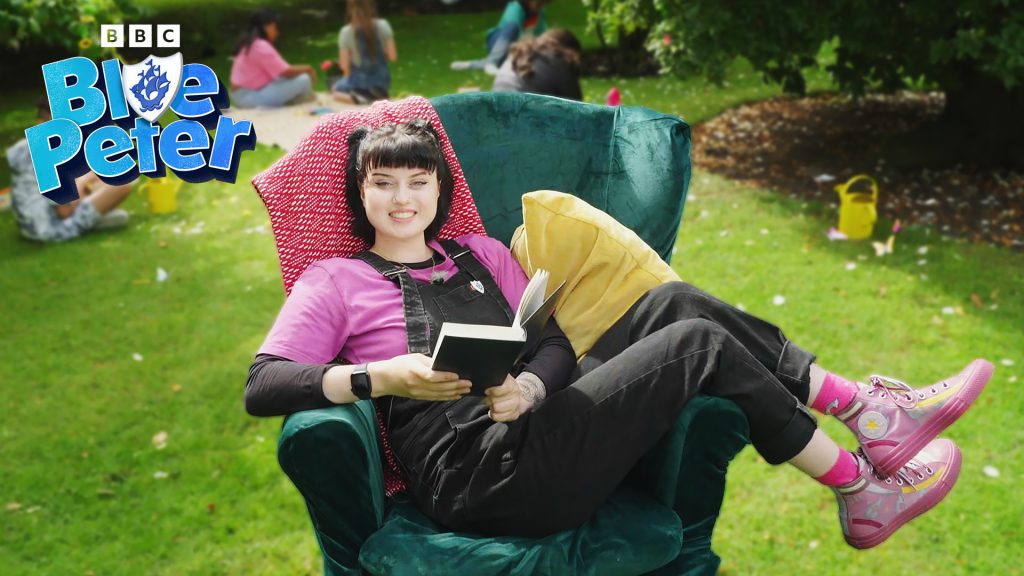 Blue Peter presenter, Abi, lies across an armchair in a green garden holding a book and smiling. She wears a pink t shirt, pink trainers and black dungarees.