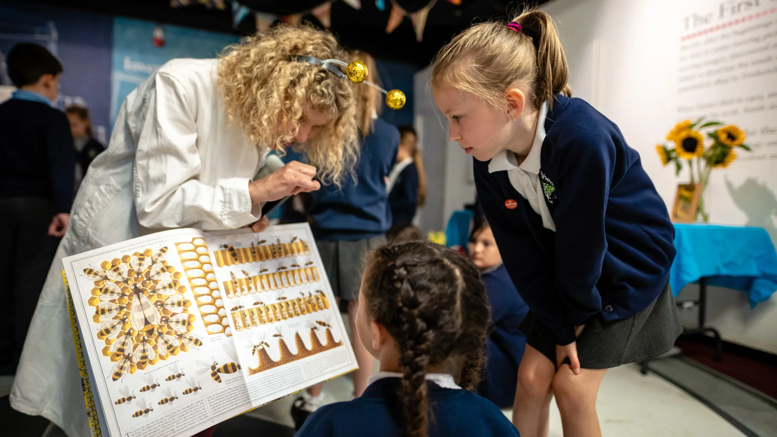 Photo of woman in a white lab coat showing two school girls an open science book with diagrams.