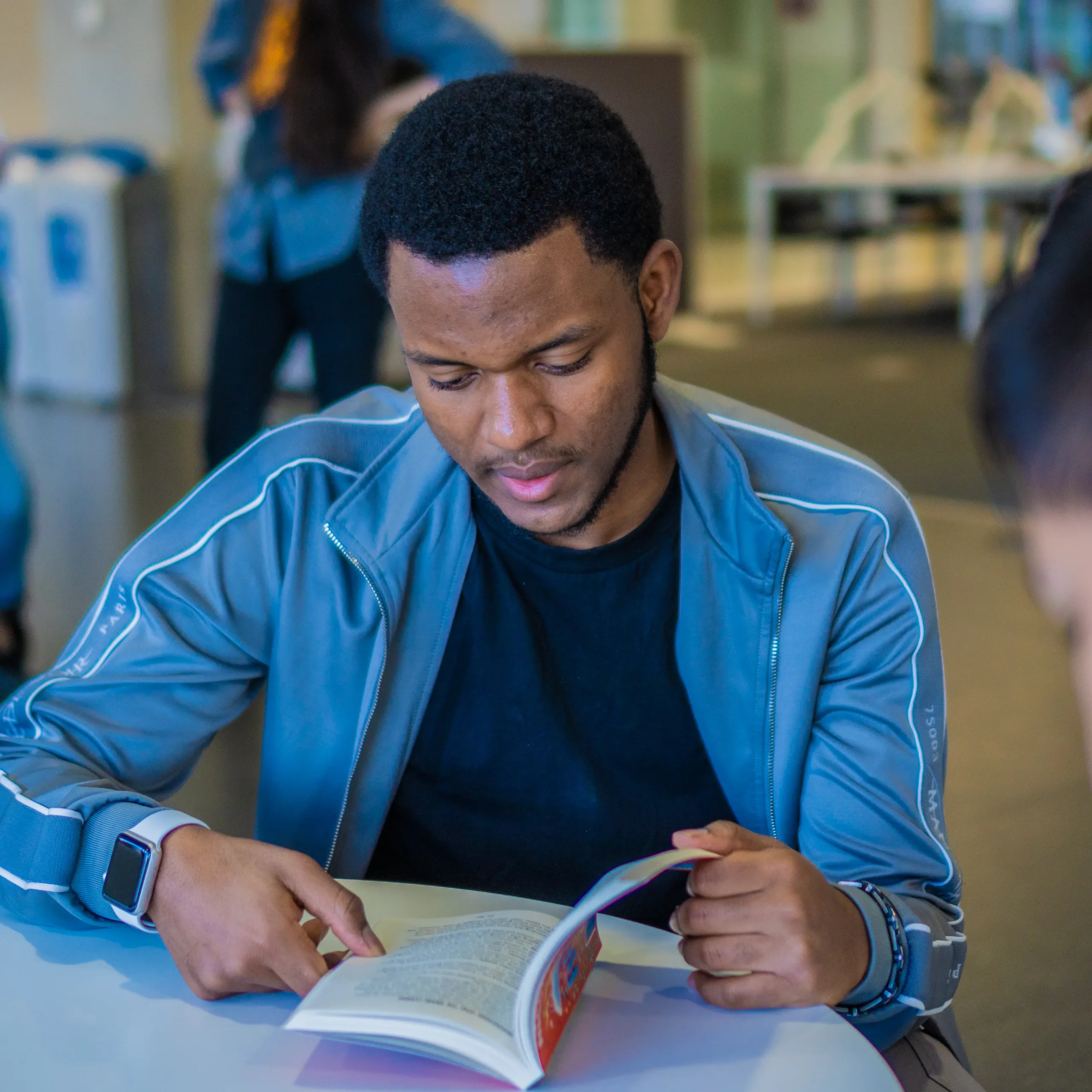 A man in a blue shirt sits at a library table reading